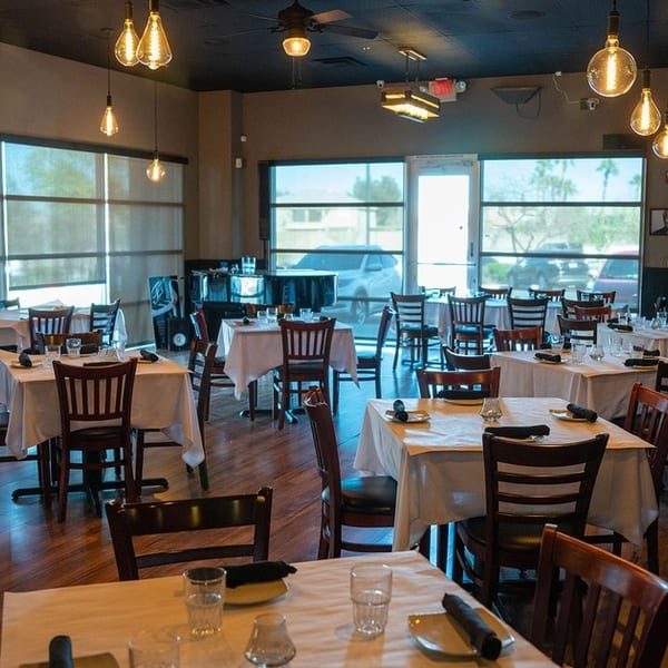 Empty restaurant dining room with set tables, dark wood chairs, and large windows.