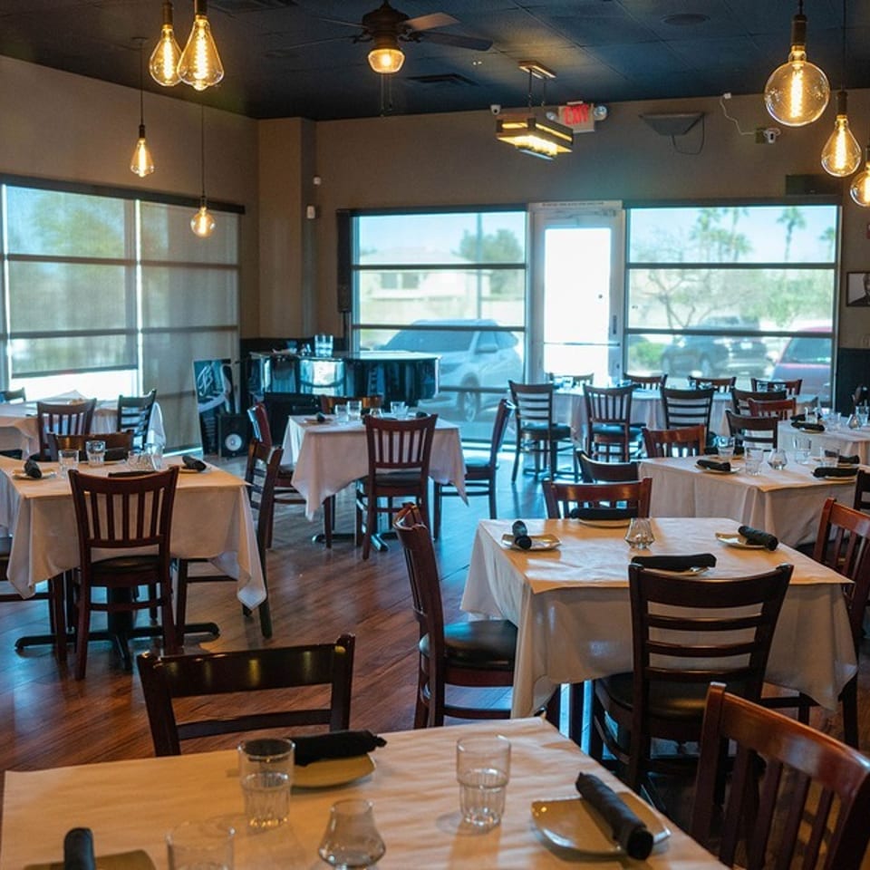 Empty restaurant dining room with set tables, dark wood chairs, and large windows.