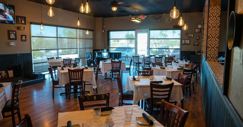 Empty restaurant dining room with set tables, dark wood chairs, and large windows.