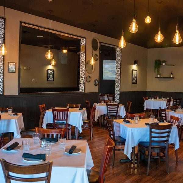 An empty restaurant dining room with tables set with white tablecloths and dark wooden chairs.