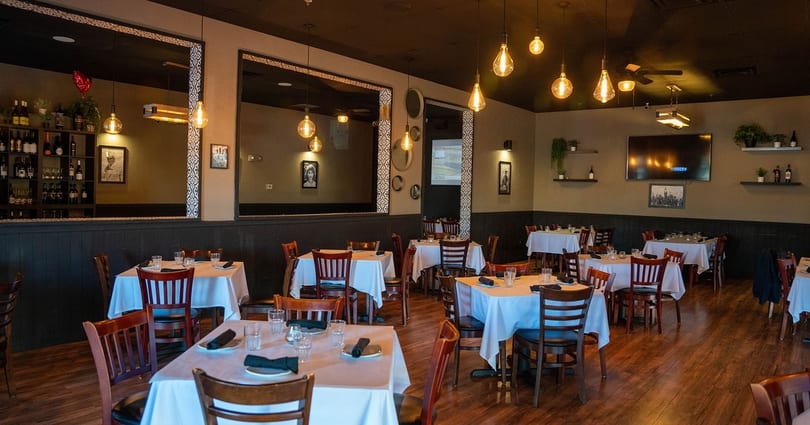 An empty restaurant dining room with tables set with white tablecloths and dark wooden chairs.