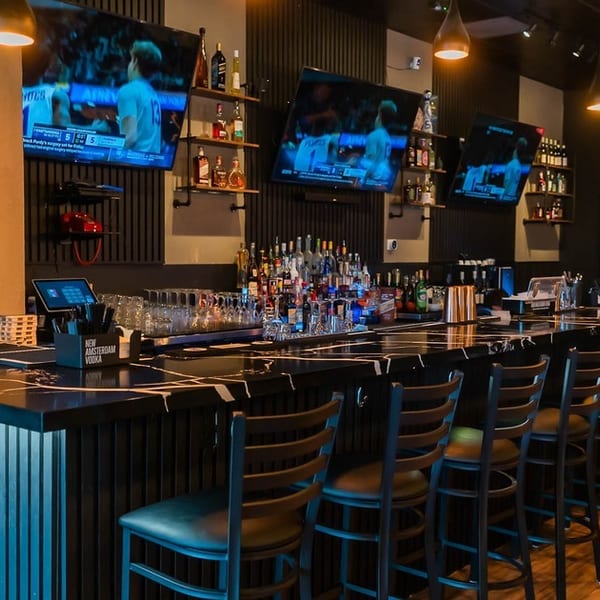 A modern bar interior with a long black marble counter, bar stools, liquor bottles, and TVs.