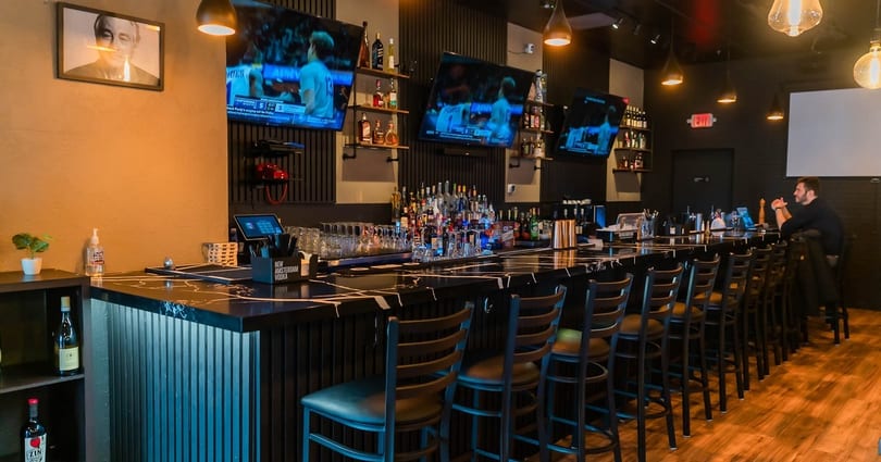 A modern bar interior with a long black marble counter, bar stools, liquor bottles, and TVs.