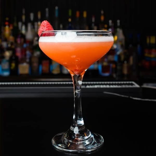 A vibrant orange cocktail with a foamy top and raspberry garnish sits on a dark bar.