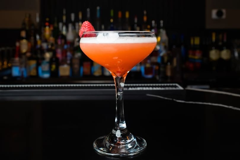 A vibrant orange cocktail with a foamy top and raspberry garnish sits on a dark bar.