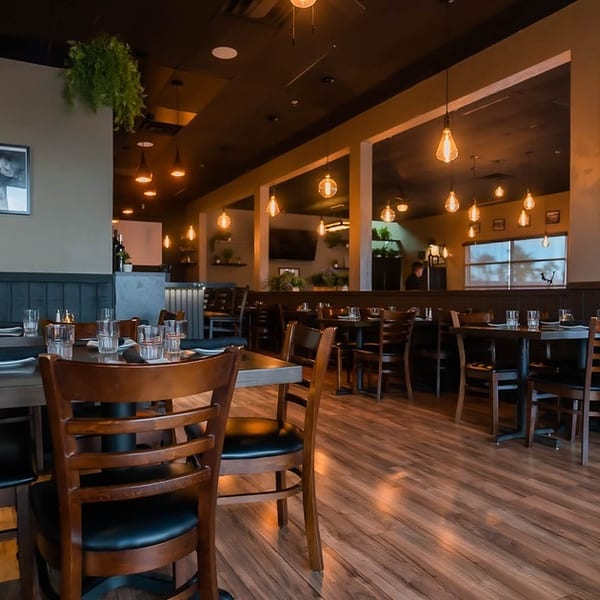 A restaurant dining room with dark wood tables, chairs, and warm hanging Edison bulb lights.