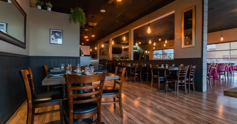 A restaurant dining room with dark wood tables, chairs, and warm hanging Edison bulb lights.