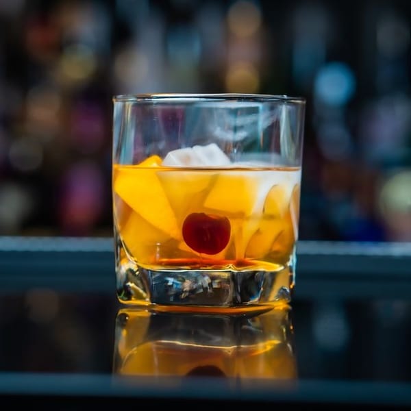 An Old Fashioned cocktail with orange slices, ice, and a cherry, on a reflective bar.