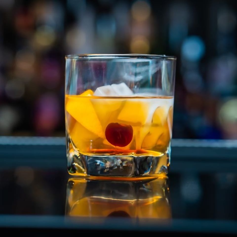 An Old Fashioned cocktail with orange slices, ice, and a cherry, on a reflective bar.