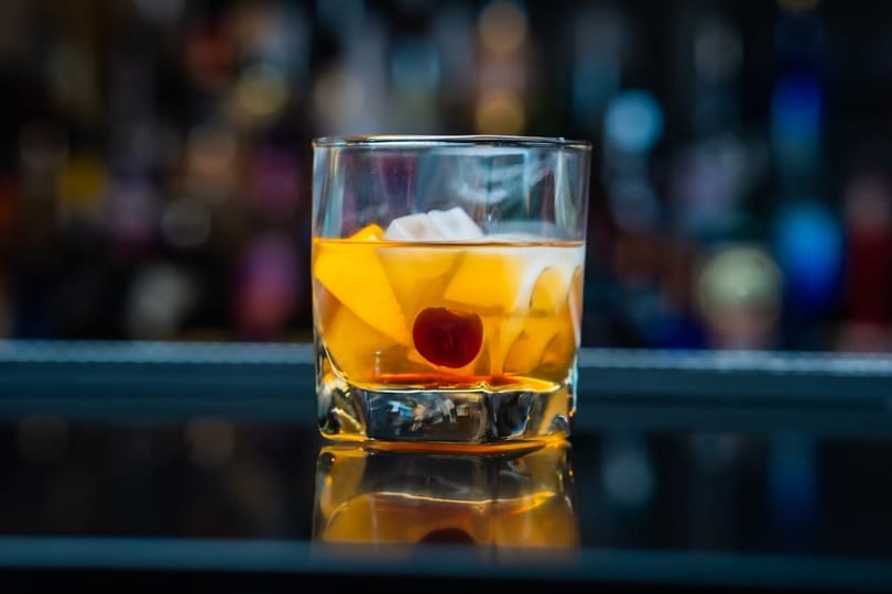An Old Fashioned cocktail with orange slices, ice, and a cherry, on a reflective bar.