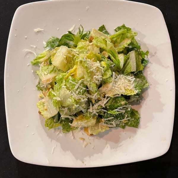 A Caesar salad with romaine lettuce, croutons, and shredded Parmesan cheese on a white square plate.