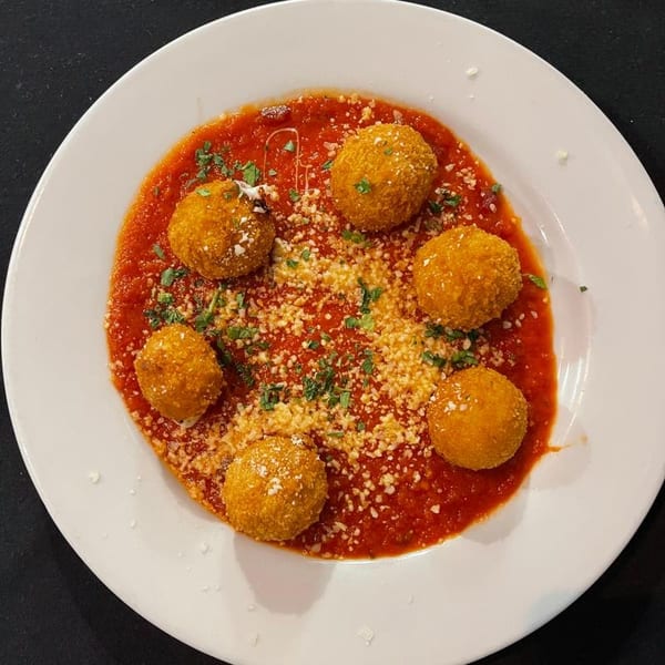 Six golden-brown arancini balls in a vibrant red marinara sauce, topped with grated cheese and parsley.