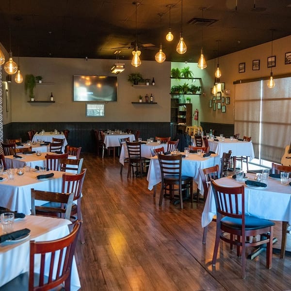 Restaurant dining room with white-clothed tables, wooden chairs, hanging Edison bulbs, and wall-mounted TVs.