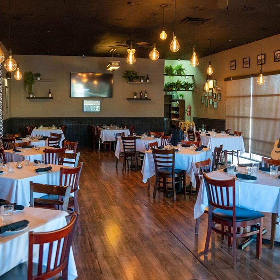 Restaurant dining room with white-clothed tables, wooden chairs, hanging Edison bulbs, and wall-mounted TVs.