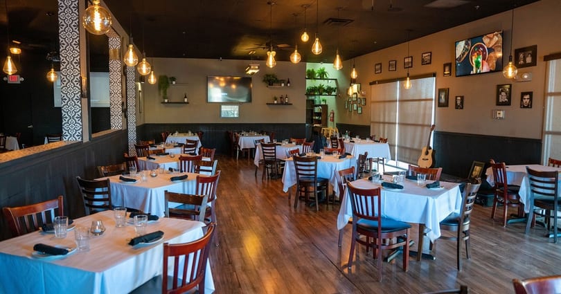 Restaurant dining room with white-clothed tables, wooden chairs, hanging Edison bulbs, and wall-mounted TVs.