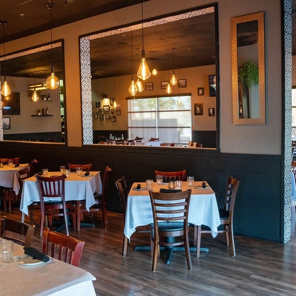 Restaurant interior with set tables, dark wood floors, large mirrors, and warm pendant lighting.