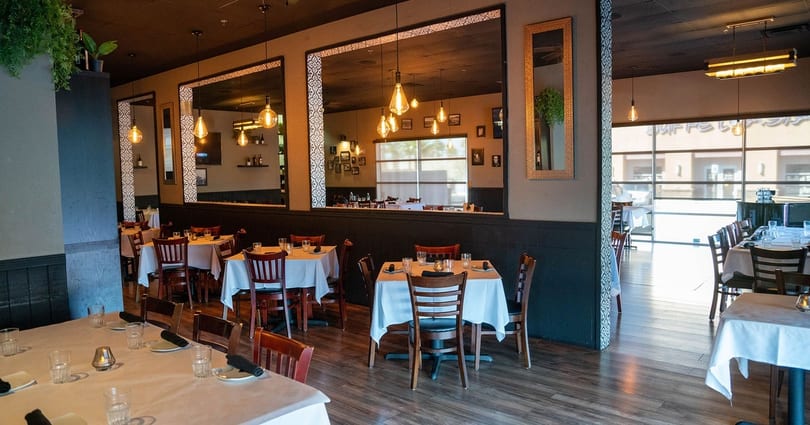 Restaurant interior with set tables, dark wood floors, large mirrors, and warm pendant lighting.