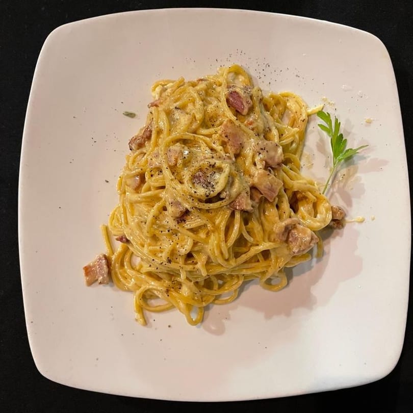 Creamy carbonara pasta with bacon bits, black pepper, and a parsley garnish on a white plate.